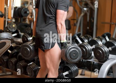 Primo piano di un maschio muscoloso non identificabile che raccoglie pesi pesanti dumbbell da rack attrezzature in palestra moderna. Foto Stock
