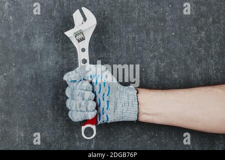Mano dell'uomo che tiene una chiave regolabile su sfondo grigio, mano dell'uomo in guanti Foto Stock