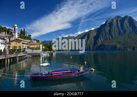 Riva di Solto, tipica cittadina italiana sul Lago d'Iseo con una chiesa e una passeggiata sulla spiaggia. Una barca blu e rossa di fronte. Monte Corna Trentapassi Foto Stock