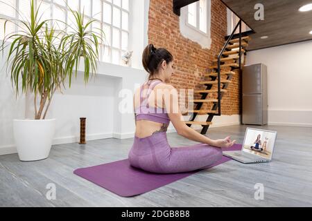 Giovane donna a casa che prende una lezione di yoga online sul suo computer portatile. Concetto di benessere e nuove tecnologie. Spazio per il testo. Foto Stock