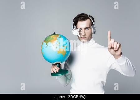 Cyborg in cuffie che tengono il globo mentre puntano verso l'alto con il dito isolato su grigio Foto Stock