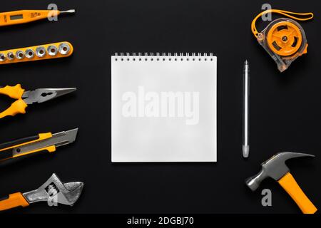 Una serie di strumenti di costruzione e un foglio bianco di carta con una penna. Sfondo nero. Scheda Labor Day, notebook al centro con spazio per il testo. Foto Stock