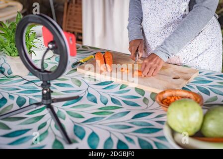 donna nera con gli occhiali insegna online con il suo telefono cellulare lezioni di cucina da casa Foto Stock