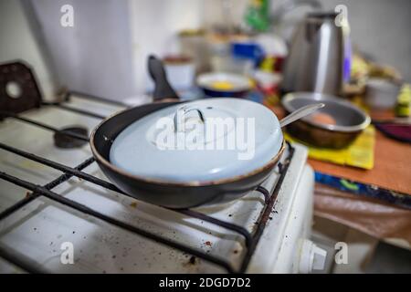 Una vecchia padella piccola coperta da un coperchio si alza su una stufa a gas sporca Foto Stock