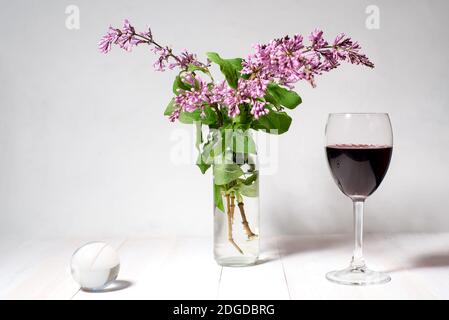 Ancora vita da un bouquet di lilla in un vaso, un bicchiere di vino e una palla trasparente su una schiena bianca Foto Stock