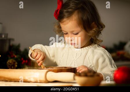 Carino piccola ragazza seria 2-4 con un arco rosso prepara i biscotti di pan di zenzero di Natale nella cucina del capodanno. Foto Stock