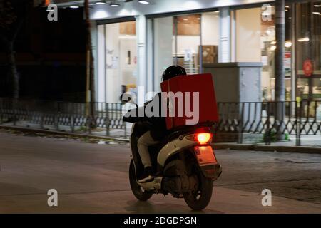 Corriere di consegna su moto senza marchio che velocizza sulla strada della città di notte. Vista illuminata di un maschio con moto da cavalcare a casco per consegnare il cibo da asporto contenuto in una scatola rossa a Salonicco, Grecia. Foto Stock