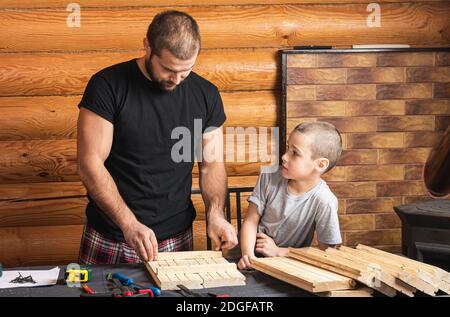 Papà e figlio stanno lavorando su un prodotto in legno, realizzando contrassegni per il fissaggio, gli attrezzi e il legno sul tavolo in officina. Concetto di formazione di carpenteria Foto Stock