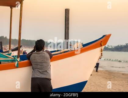 Il giovane indiano ammira il tramonto sulla spiaggia sabbiosa che si affaccia su una barca da pesca Foto Stock