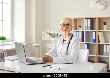 Medico professionale senior positivo in uniforme seduto con il computer portatile in ufficio medico Foto Stock