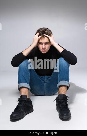 Elegante ragazzo in posa in studio seduto sul pavimento in pantaloni in denim e felpa nera. Ritratto di studente maschile pieno su sfondo bianco Foto Stock