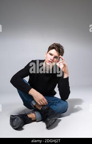 Elegante ragazzo in posa in studio seduto sul pavimento in pantaloni in denim e felpa nera. Ritratto di studente maschile pieno su sfondo bianco Foto Stock