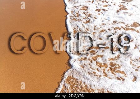 La parola COVID-19 è scritta come testo sullo sfondo di spiaggia di sabbia e schiuma onda Foto Stock