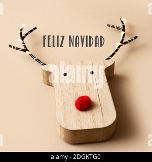una divertente testa di renna in legno, con un naso rosso, e il testo buon natale in spagnolo su uno sfondo marrone chiaro Foto Stock