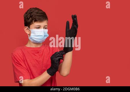 Un ragazzo adolescente in una maschera medica mette su guanti protettivi. Su sfondo rosso. Concetto di coronavirus e pandemia. Foto Stock