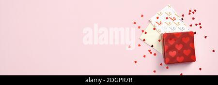 Scatola regalo rossa, biglietto d'auguri e busta su sfondo rosa pastello con confetti a forma di cuore. Modello banner giorno di San Valentino. Vista dall'alto con spin. Copia Foto Stock