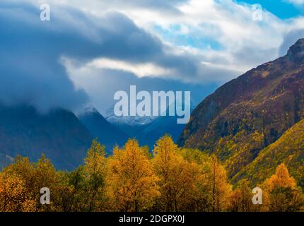 Valle con alberi in colori autunnali, suggestivo paesaggio di nuvole e pendii di montagna e picchi in lontananza. Foto Stock