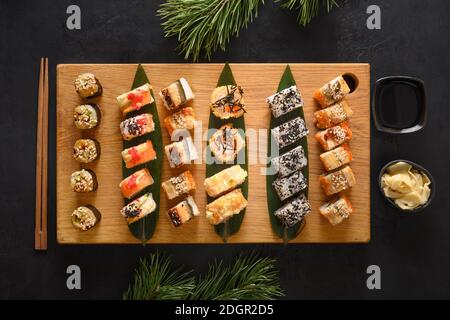 Set di sushi natalizi servito su tagliere di legno con decorazioni natalizie su sfondo nero. Vista dall'alto. Stile di posa piatto. Foto Stock