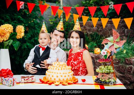 Tema compleanno bambini. Famiglia di tre persone caucasiche seduta nel cortile della casa ad una festa decorato tavolo in divertente Foto Stock