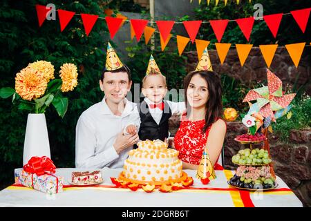 Tema compleanno bambini. Famiglia di tre persone caucasiche seduta nel cortile della casa ad una festa decorato tavolo in divertente Foto Stock