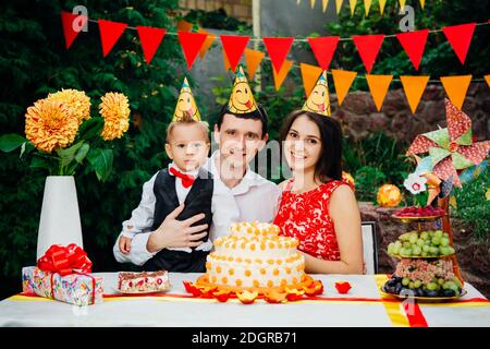 Tema compleanno bambini. Famiglia di tre persone caucasiche seduta nel cortile della casa ad una festa decorato tavolo in divertente Foto Stock