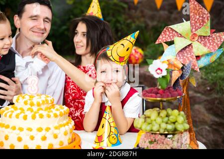 Ragazza festa di compleanno dei bambini seduto a un tavolo vicino a una torta in un cappello festivo e sorridente Foto Stock