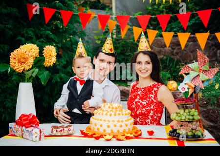 Tema compleanno bambini. Famiglia di tre persone caucasiche seduta nel cortile della casa ad una festa decorato tavolo in divertente Foto Stock