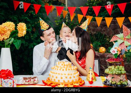 Tema compleanno bambini. Famiglia di tre persone caucasiche seduta nel cortile della casa ad una festa decorato tavolo in divertente Foto Stock