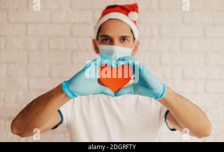 Giovane medico maschile a Santa Hat, maschera medica viso, guanti protettivi tenere il cuore rosso nelle mani. Felice uomo chirurgo in maschera protettiva viso mostra cuore sig Foto Stock