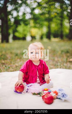 Bambina caucasica etnia bionda un anno da nascita siede su una plaid su erba verde nel parcheggiare e tenere in mano Foto Stock