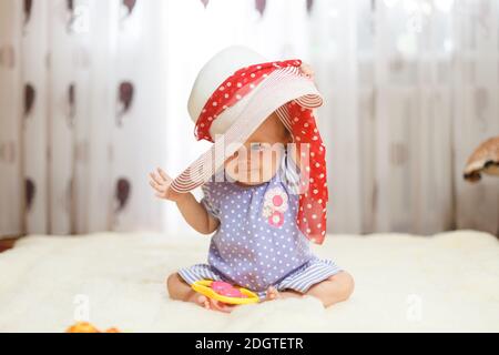 Una bambina caucasica un bambino si siede a casa sul pavimento su un tappeto leggero. Sulla sua testa è un grande cappello estivo che Foto Stock