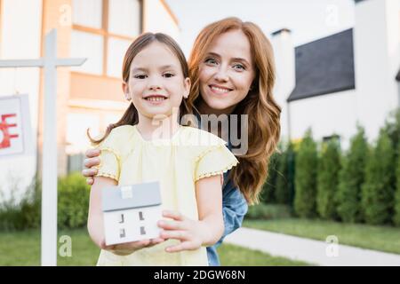 Redhead madre abbracciare figlia con statuetta casa mentre si guarda fotocamera con case sfocate sullo sfondo Foto Stock