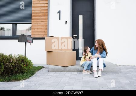 Felice madre e figlia che si guardano mentre si siedono vicino alle scatole di cartone sulla porta d'ingresso Foto Stock