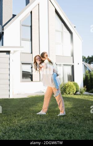 Madre sorridente piggybacking figlia vicino a casa moderna Foto Stock