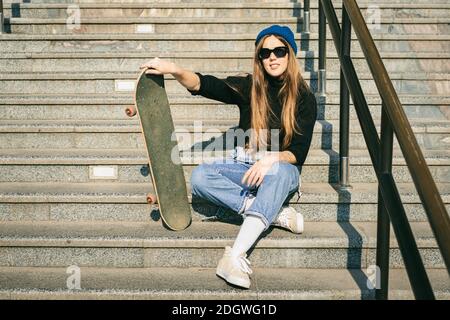 Donna elegantemente vestita con jumpsuit in denim blu in posa con skateboard. Via foto. Ritratto di ragazza che tiene skateboard. Massima vita Foto Stock