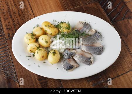 Pesce di aringa con palline di patate giovani su un piatto bianco. Sfondo di legno. Foto Stock