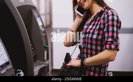 Giovane cute ragazza felice sta tirando fuori il portafoglio mentre prendendo sul telefono che si prepara ad usare ATM. Foto Stock