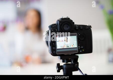 Primo piano di una fotocamera che scatta foto dei giovani bella ragazza carina che è seduto al tavolo da cucina con un computer portatile su di esso e mostrando profumi e. Foto Stock