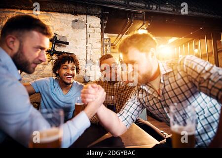 Gruppo di gioiosi felici uomini che si divertono nel pub. Due amici lottano con le braccia mentre altri sostengono. Foto Stock