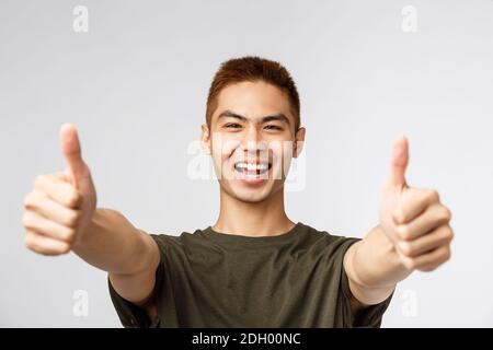 Persone, espressioni diverse e concetto di stile di vita. Entusiasta felice uomo eccitato, ragazzo asiatico mostra pollici-up e sorridente, sayin Foto Stock