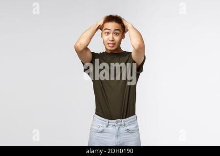 Ritratto di giovane studente asiatico alarmato di sesso maschile ha avuto problemi, tenendo le mani sulla testa e fissando fotocamera indecisiva, non so cosa Foto Stock