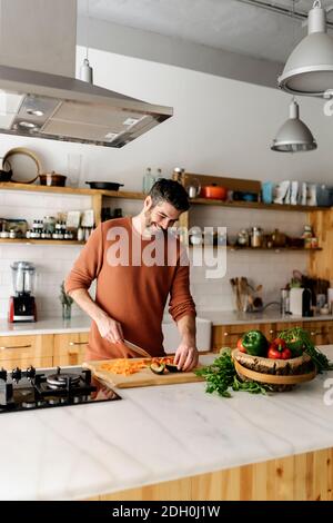 Foto di stock di un uomo di mezza età che tiene un coltello e affettare alcune verdure in cucina. Sta guardando attraverso una finestra e sorridendo. Foto Stock