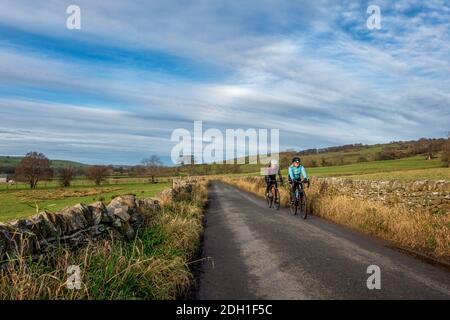 Due donne socializzano, ciclano e si tengono in forma - cavalcando in bici da strada attraverso la campagna con un alone in cielo, Ilkley, West Yorkshire, Inghilterra, Regno Unito Foto Stock