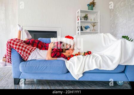 Vista laterale della coppia felice che si trova sul divano o sul divano con cappelli santa e pigiami. Giovane uomo e donna si coccolano e si guardano con tenerne Foto Stock