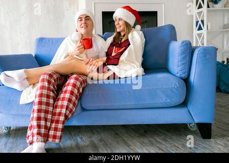 Natale, casa, amore concetto. Ritratto di giovane coppia in cappelli di Santa, coperto di plaid bianco che tiene coppe rosse, abbracciando e sorridendo mentre si siede a h. Foto Stock