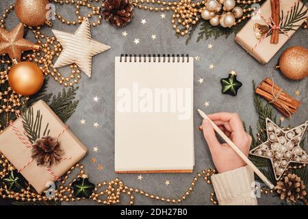 Blocco note e scrittura mano femminile con decorazioni natalizie in colori verde e oro su sfondo grigio in cemento, copyspace. Piano di Natale e Capodanno Foto Stock