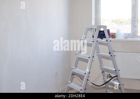 Scala pieghevole in alluminio in una stanza con pareti in intonaco bianco e finestra. Ristrutturazione dell'alloggio in una nuova casa. Spazio di copia. Foto Stock