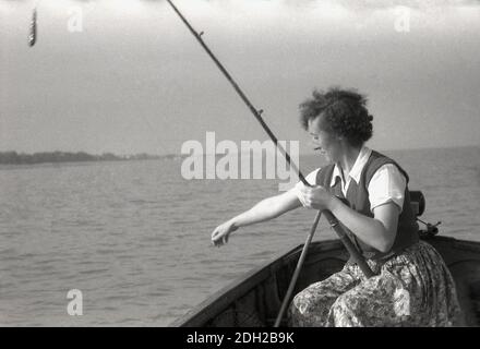 Anni 1950 storico in mare in una barca di legno una signora che indossa una gonna a motivi floreali un cardigan di lana a manica corta e superiore pesca seduto con una