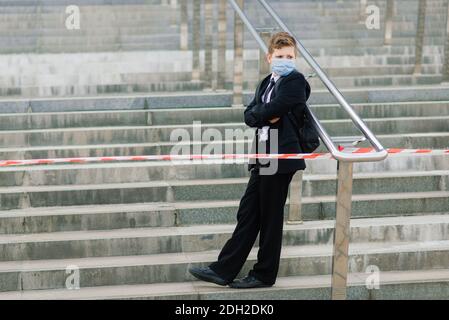 Scolaro esce dalla scuola indossando una maschera protettiva nel città Foto Stock