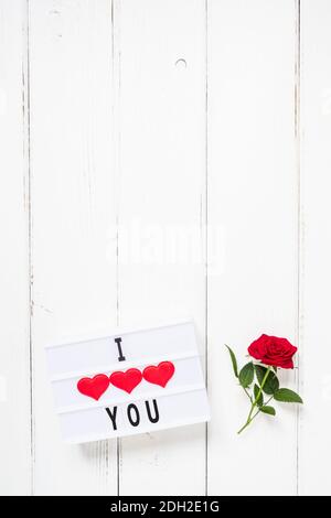 San Valentino, amore, romanticismo concetto. Fiori di rosa rosso fresco e testo TI AMO sulla scatola luminosa su sfondo di legno bianco. Biglietto d'auguri. Disposizione piatta, parte superiore Foto Stock
